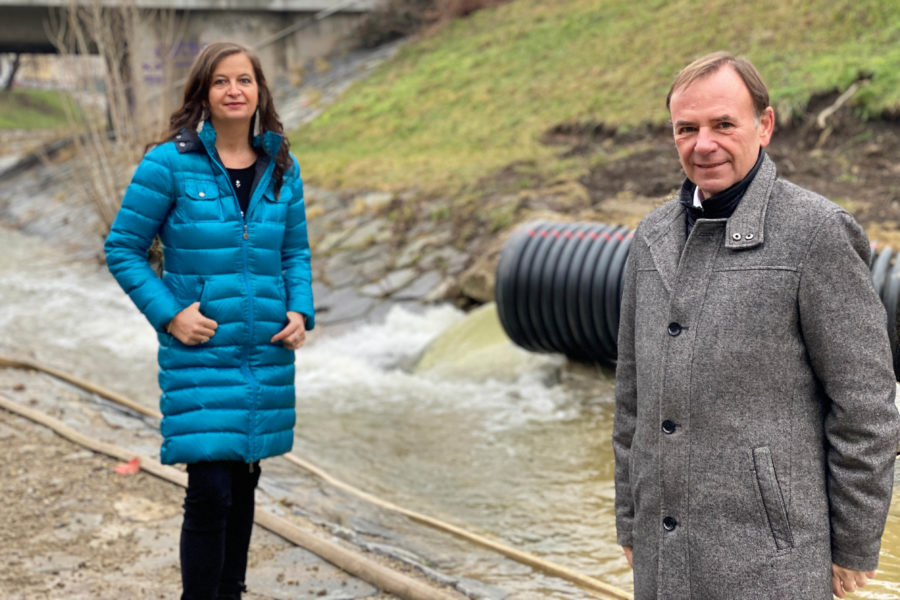 Ulli Sima und Gerald Bischof stehen auf der Baustelle der Liesingbachrenaturierung direkt neben dem Bach. Im Hintergrund sieht man den umgeleiteten Bach aus einem großen schwarzen Rohr in den hart verbauten Liesingbach reinfließen