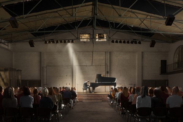 In einer großen Halle sieht man auf der Bühne einen Klavierflügen mit Pianisten und davor das Publikum