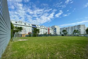 Fußballplatz mit grüner Wiese, blauem HImmel mit weißen Wolken, im Hintergrund ein Mehrfamilienhaus