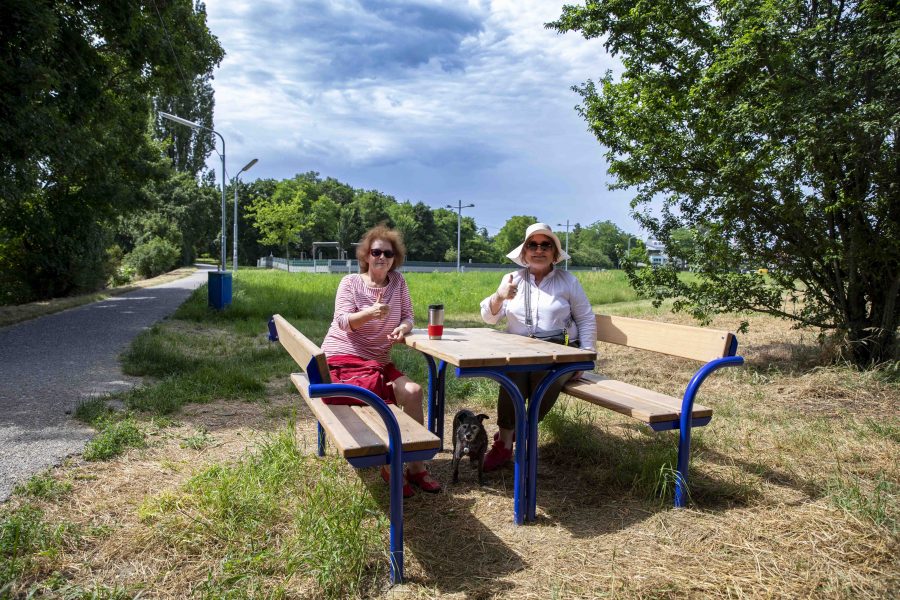 Vordergrund: Sitz-Bankkombination mit 2 Damen und einem Hund, im HIntergrund ist der Himmel und Bäume, sowie eine Hundezone zu sehen.
