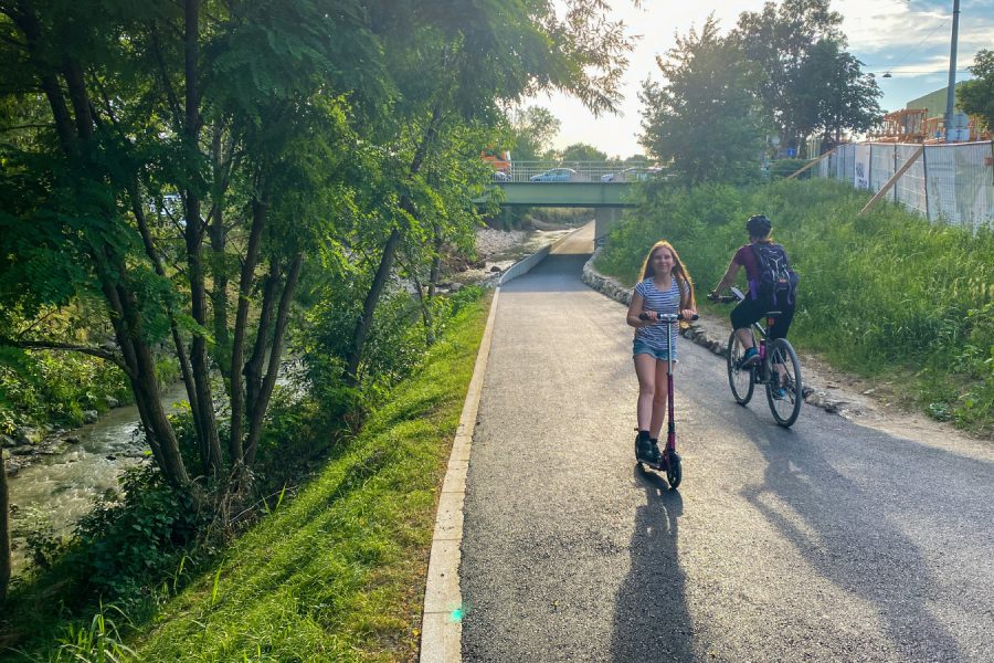 links Baume und Wiese, dann eine Asphaltweg, Kind kommt mit Roller entgegen, Radfahrer fährt weg, im Hintergrund eine Brücke